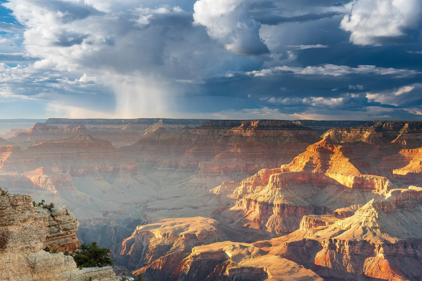 A rain shower drifting over the layered red cliffs of the Grand Canyon at sunset